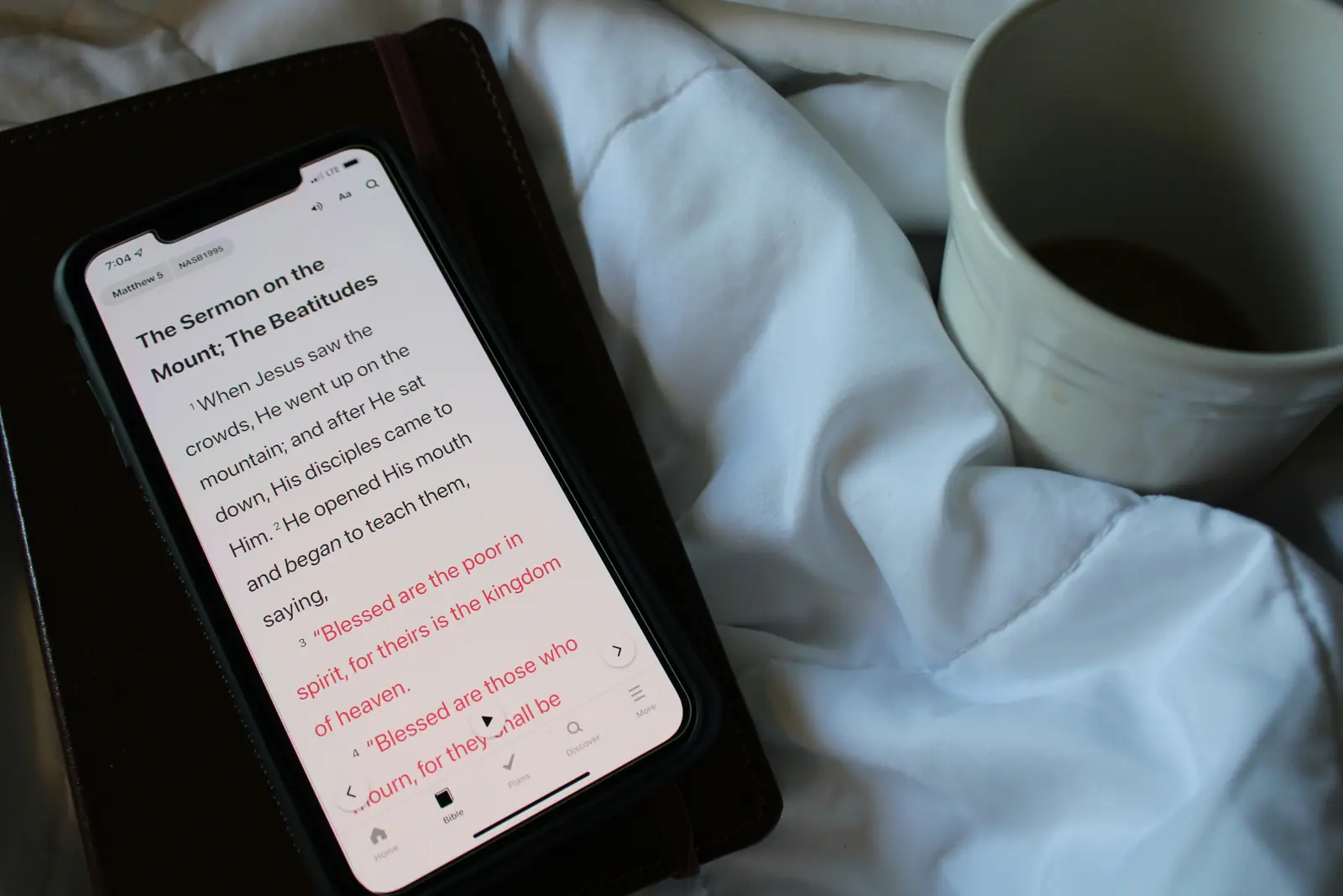 Phone open to a Bible reading app on a leather notebook next to a mug of coffee on white bedding.